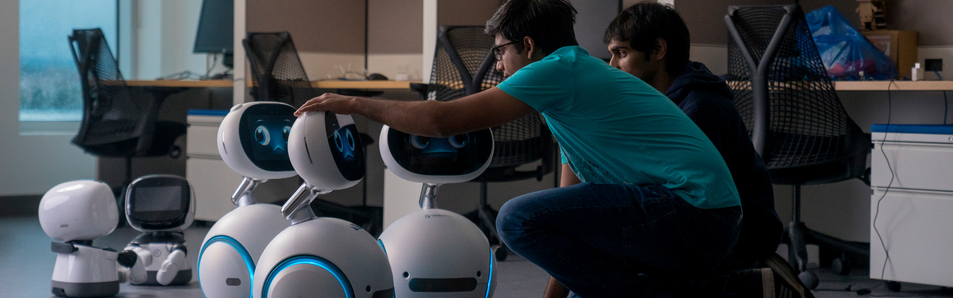 Two student kneel down, looking at three small round robots