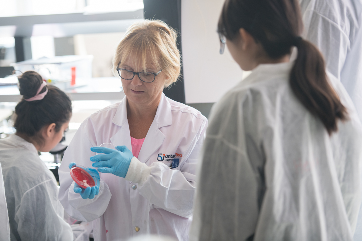academics image showing lab instructor in a white coat and blue gloves holding up a petri dish for students during a lab demonstration