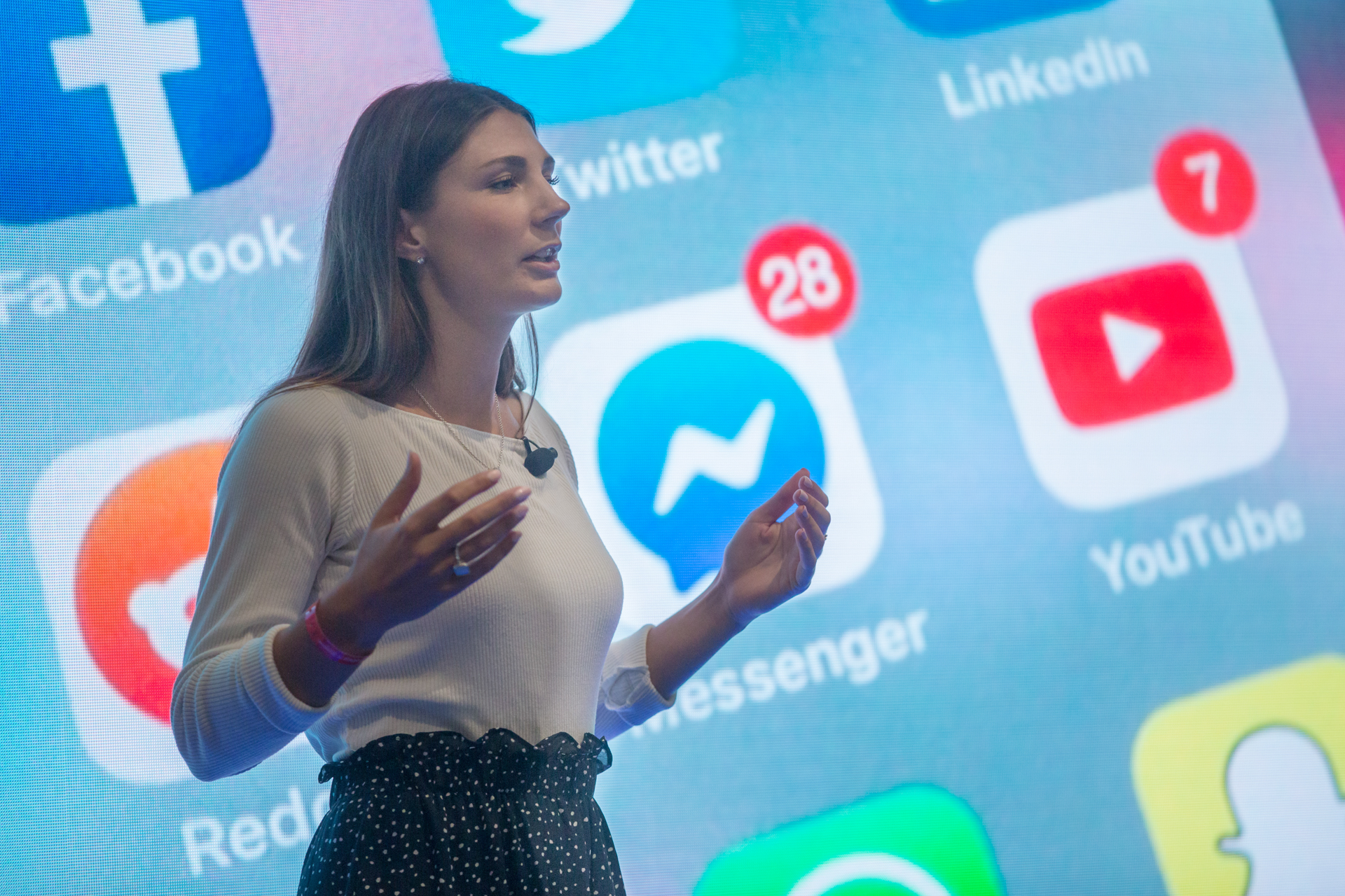A woman stands on stage, the background shows multiple social media logos.