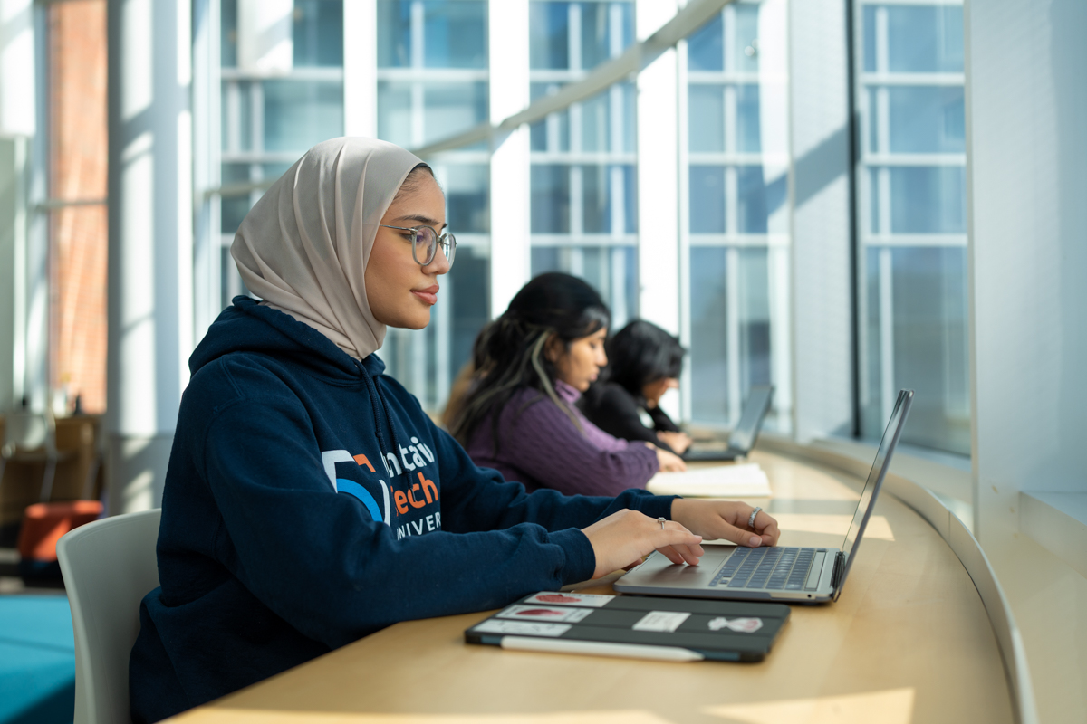 An Ontario Tech student sits in front of a sunny window in the Library, working on their laptop.
