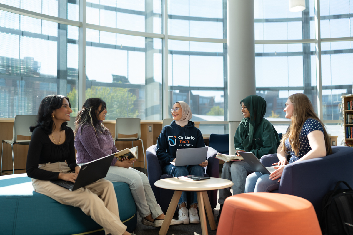 A group of Ontario Tech students, laptops on hand and ready to learn, happily chat in the Library Fireside Reading Room.