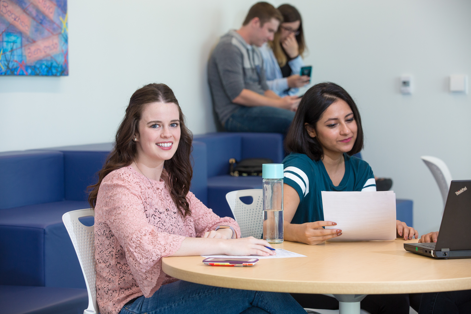 Two students sit at a table, one of them is smiling and looking at the camera while the other studies.