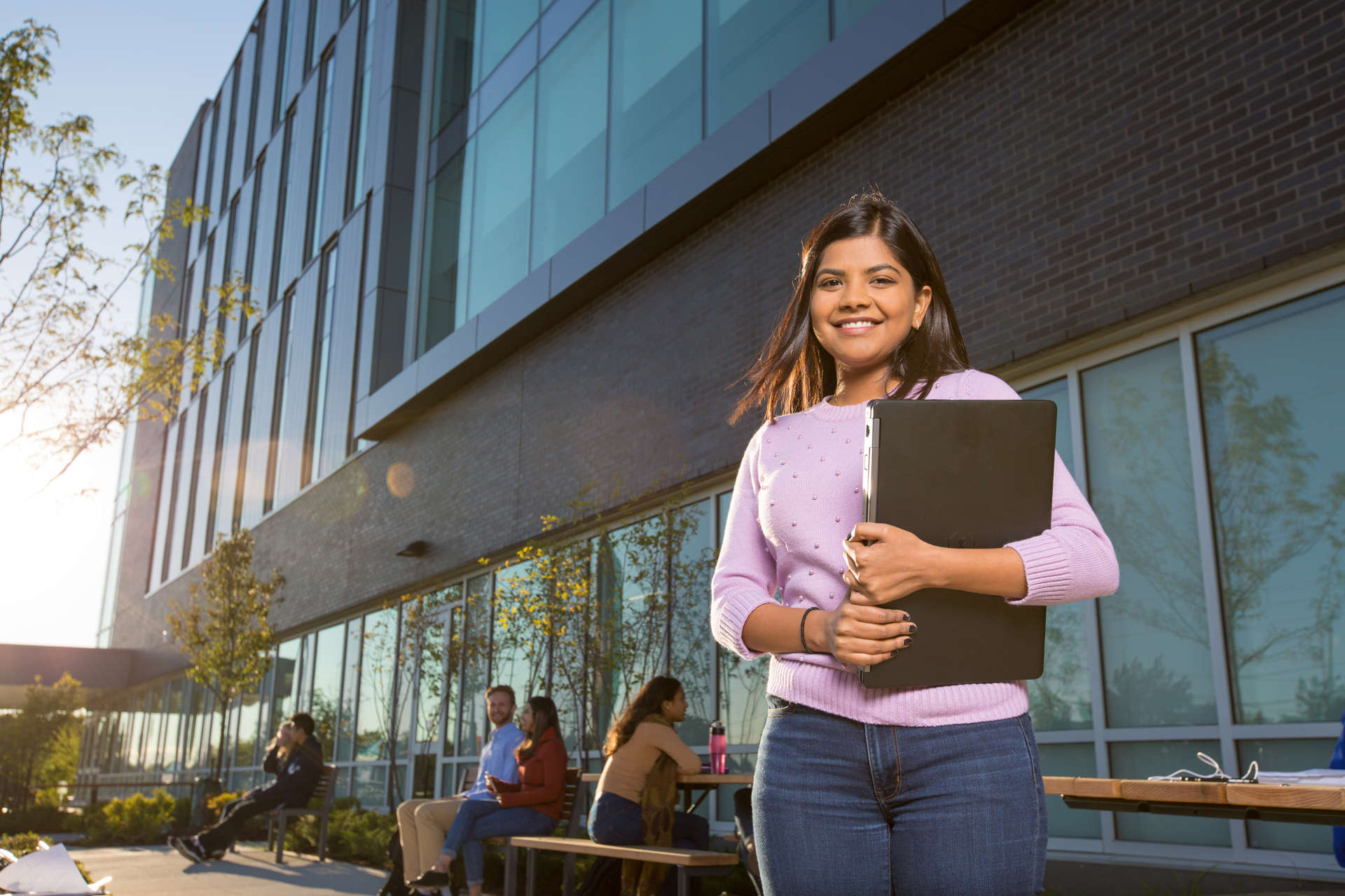 A student in a soft pink cardigan stands in front of a glass-walled building. She holds a closed laptop in her arms and is smiling. Other students are seated at picnic tables behind her, chatting and working.